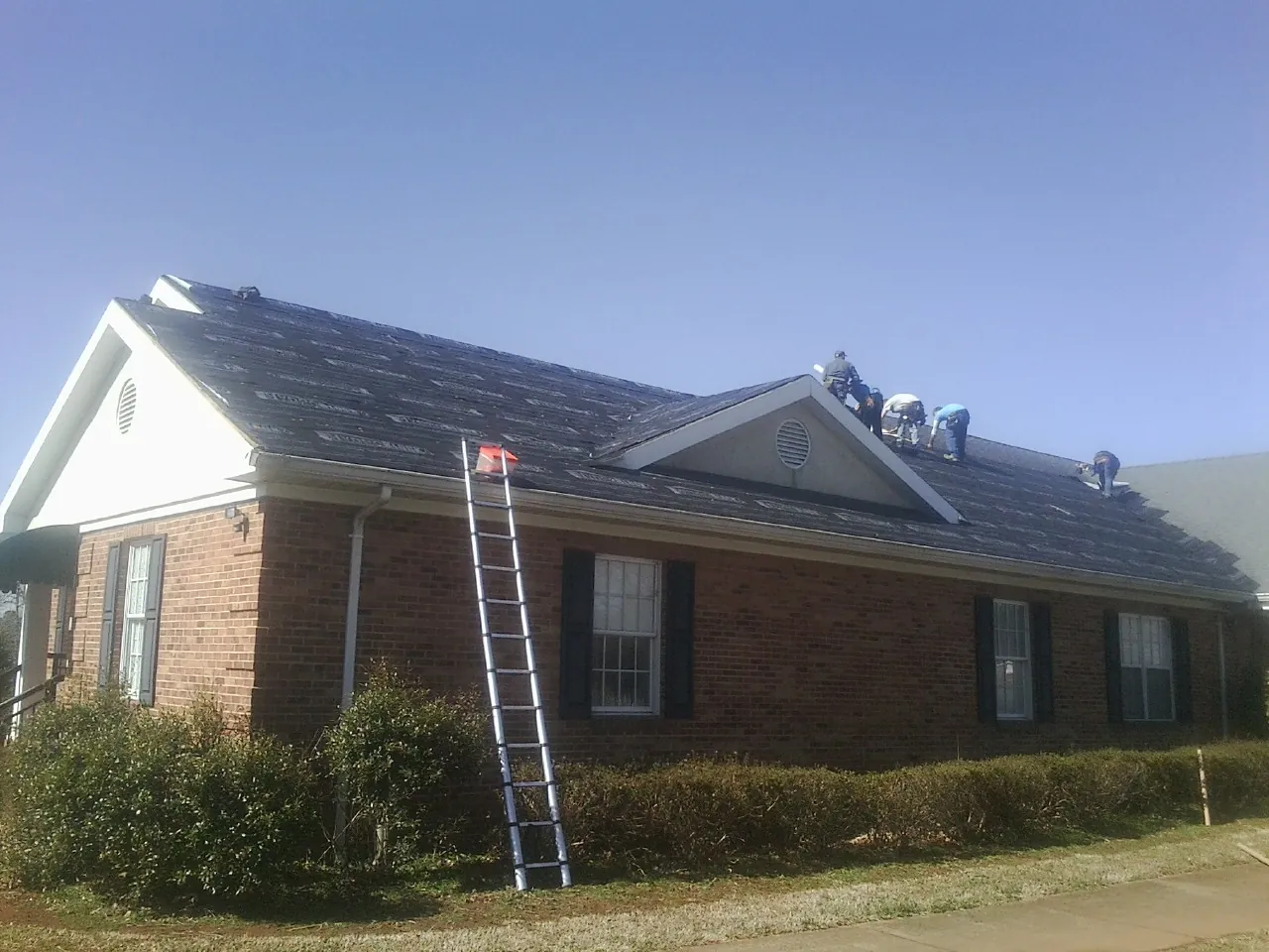 Crew tearing shingles off a brick ranch roof with a ladder set — Emergency Roof Repair work by our crew in Heath Springs