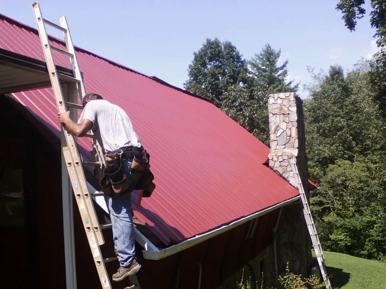 Roofer on a ladder beside a red metal roof and stone chimney — Soffit & Fascia Repair by Roofing Brothers in Heath Springs